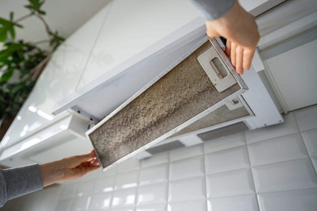 Two hands drawing a grease-loaded metal filter out from under a range hood