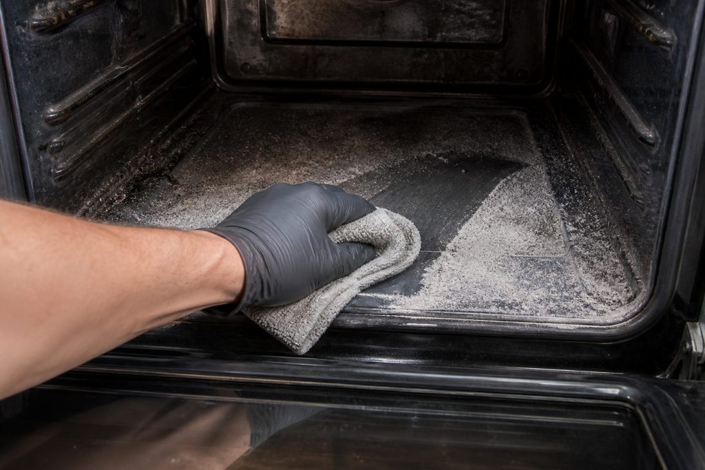 A gloved hand wiping a heavily soiled oven cavity blackened across its floor and back wall