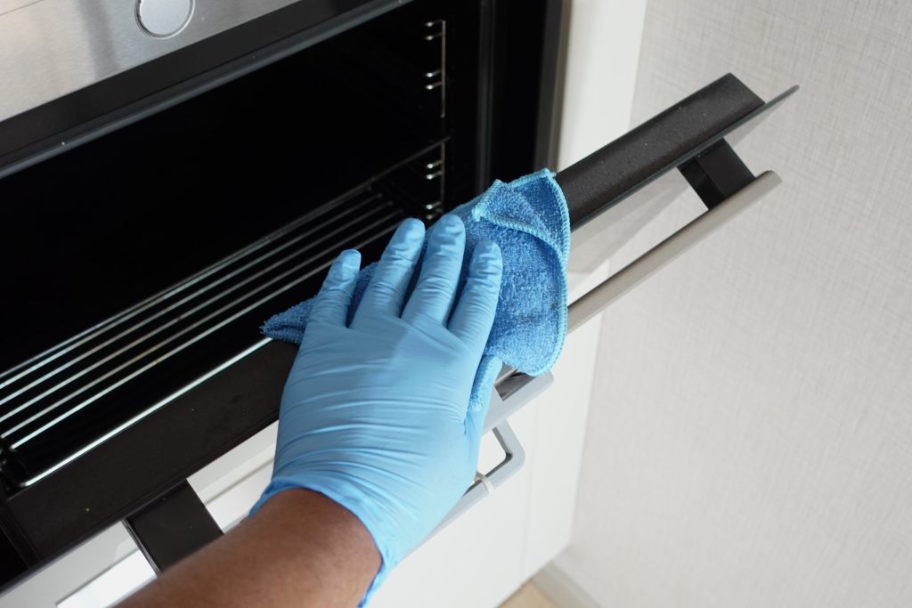 A gloved hand wiping the glass and trim along the front edge of a built-in oven door