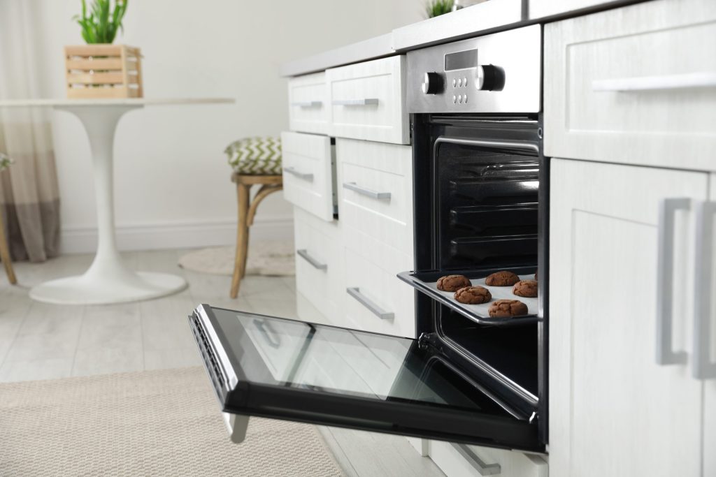 A tray of muffins resting on the open door of a built-in oven in a white kitchen