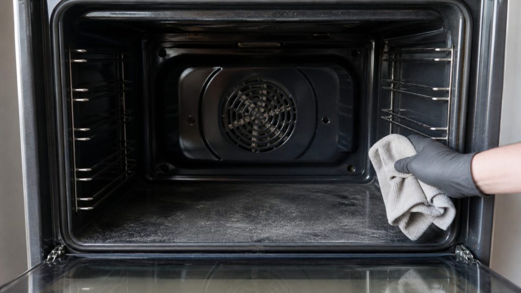 A gloved hand wiping the inside of an oven cavity with a cloth, the convection fan cover behind