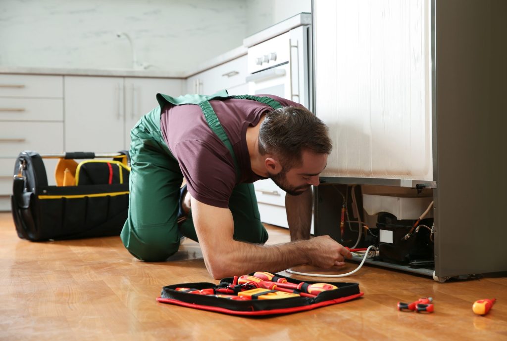 A technician lying on the floor working behind a refrigerator with a tool roll open beside him