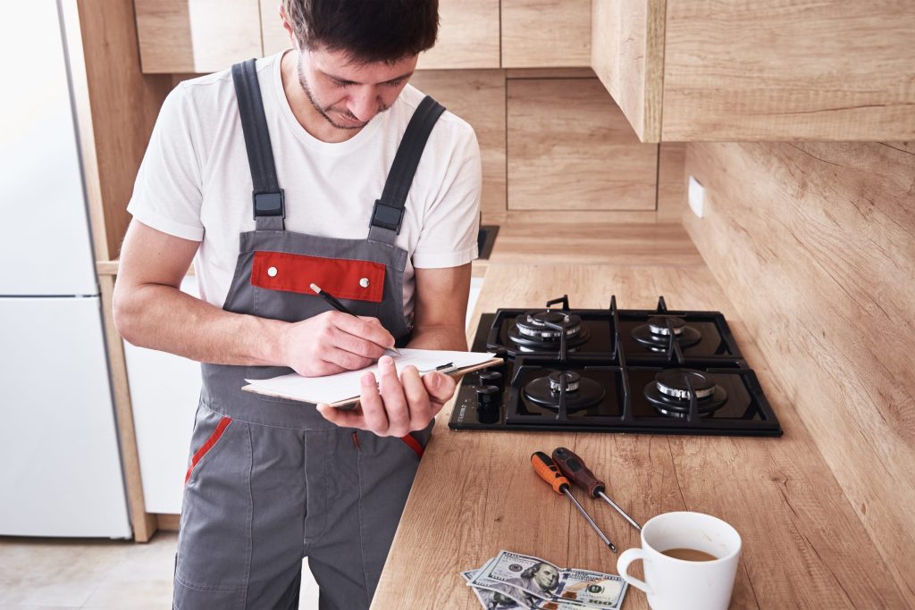 A technician standing in a kitchen writing on a pad beside a cooktop with his tools laid out
