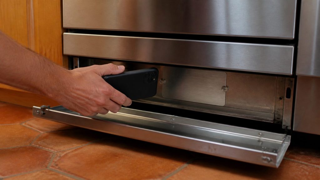 A hand holding a phone low behind the open kick panel of a stainless cooking range, photographing a small blank metal data plate riveted to the frame.