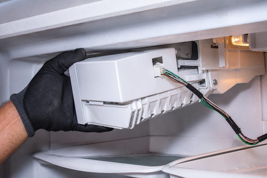 Gloved hands drawing an ice maker module out of a freezer compartment with its wiring loom still attached
