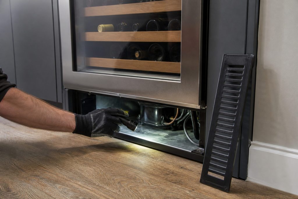 A gloved hand reaching into the base of a wine cabinet with its front grille lifted off and set on the floor
