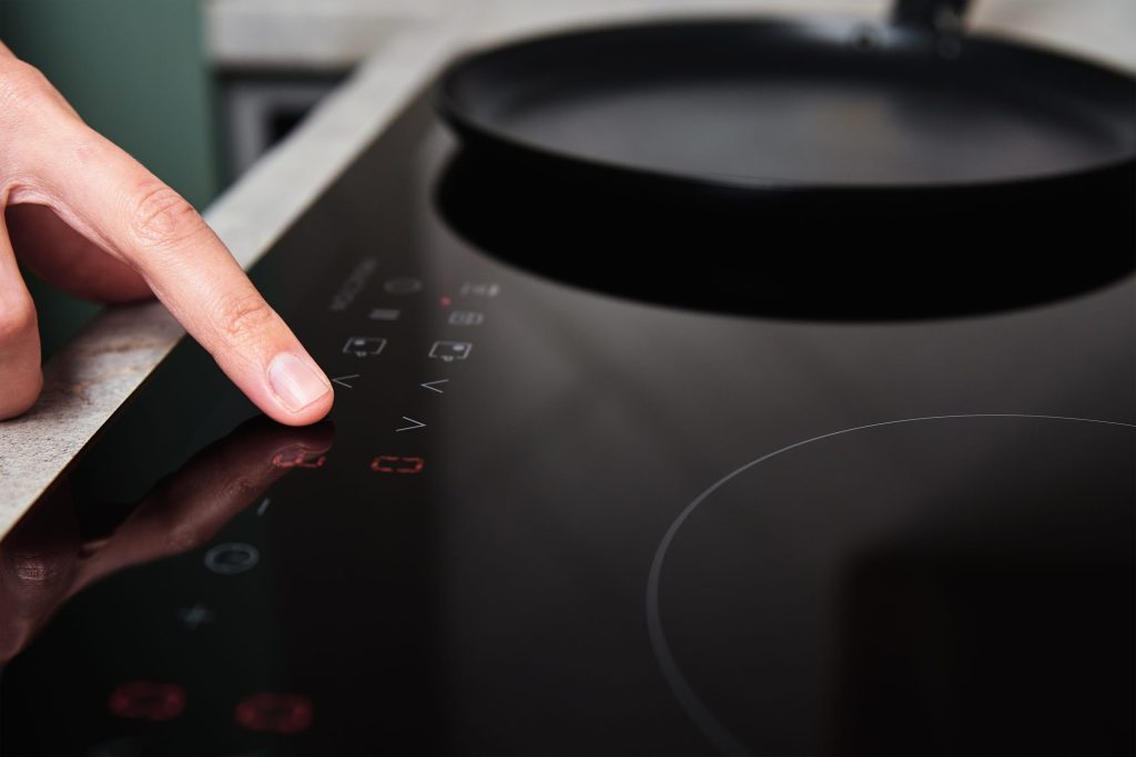 A finger pressing a touch control on a dark induction cooktop beside a pan