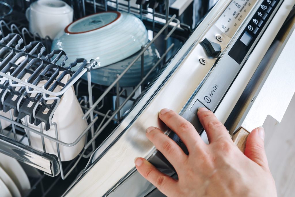 Two hands at the control strip along the top edge of an open integrated dishwasher door