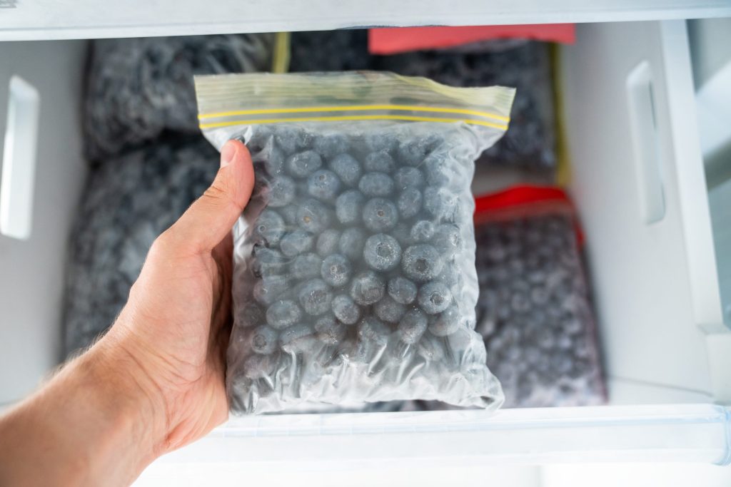 A hand holding a bag of frozen berries taken from a freezer drawer packed with other bags