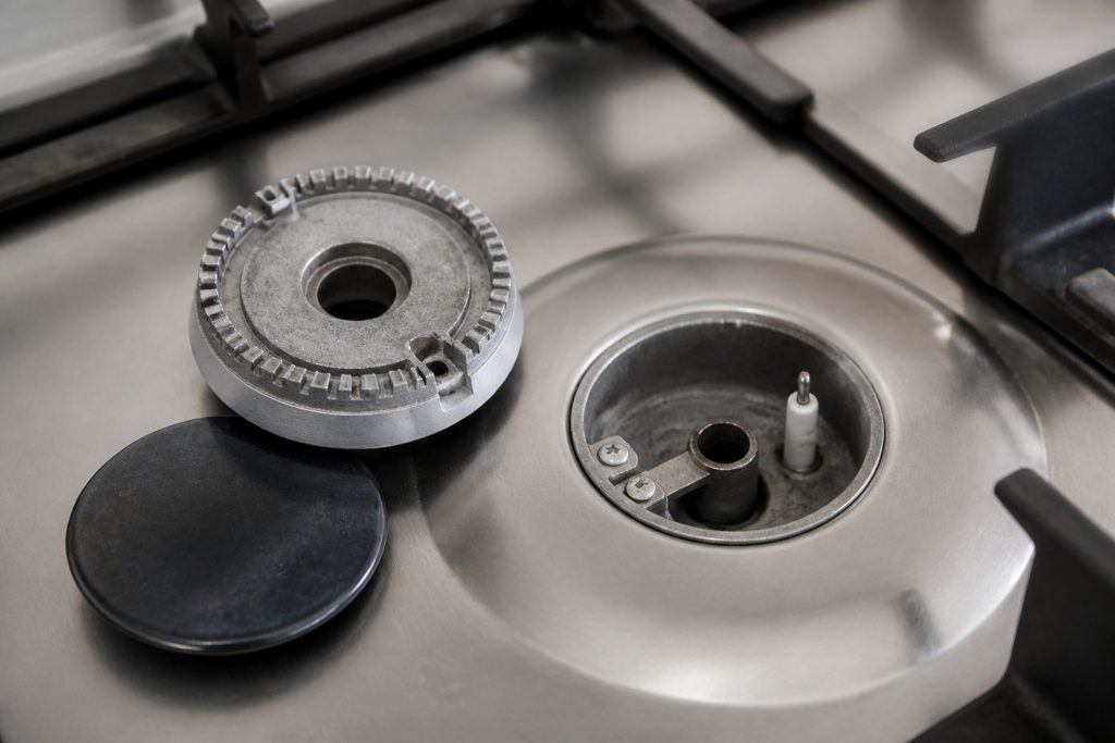 A gas burner with its cap and crown lifted off and set on the hob beside the open burner base