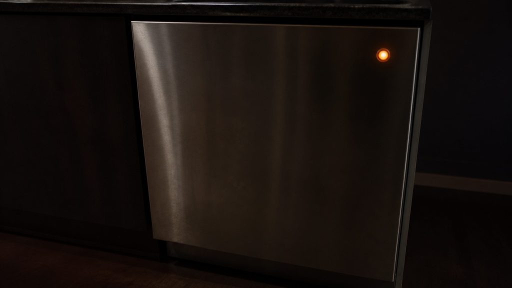 The dark front of a closed dishwasher in a dim kitchen with one small orange indicator lit