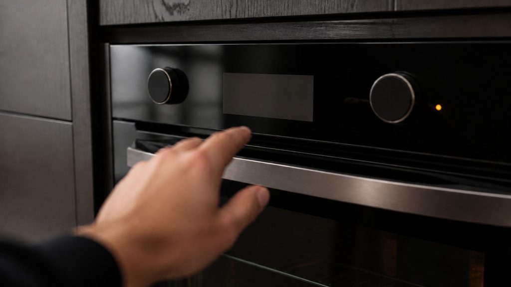 A close view of an unbranded oven control fascia in a dark kitchen with a small amber indicator lit and nothing legible on the display, a hand reaching toward it out of focus in the foreground.