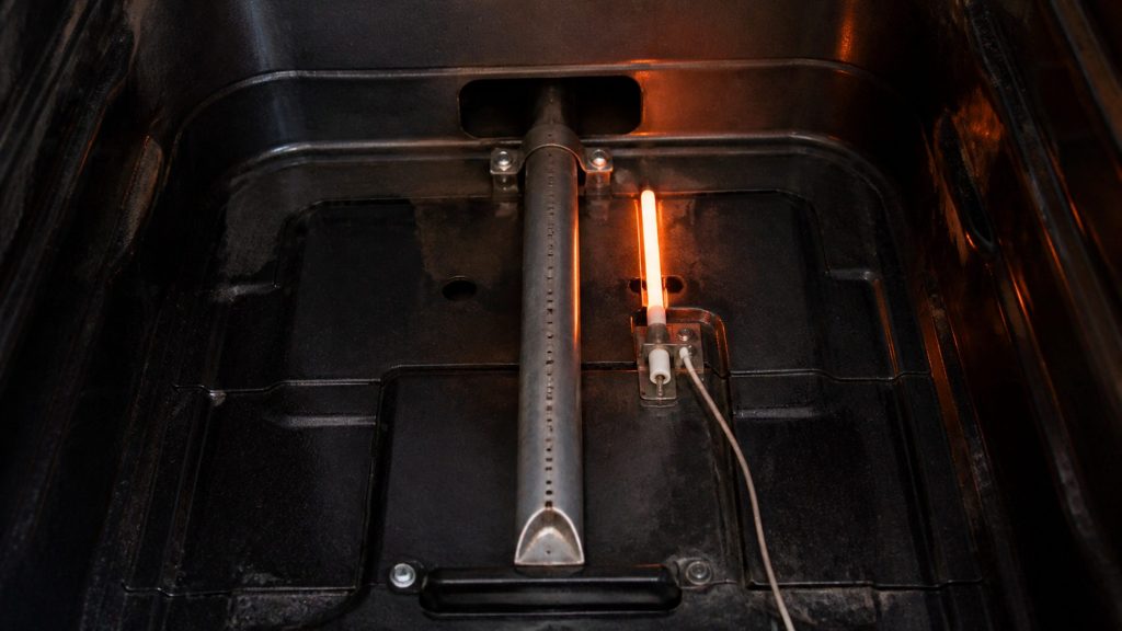 Looking into the bottom of a gas oven cavity with the floor panel lifted out, a slim ceramic igniter glowing bright orange beside the bake burner tube, no flame yet.