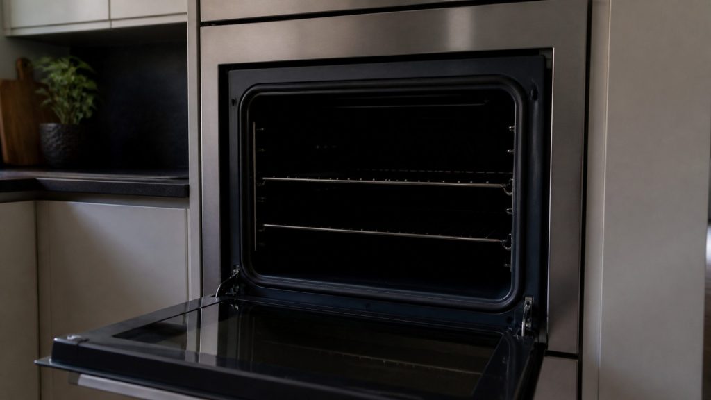 An open built-in oven in a dim kitchen with the interior completely dark, the empty racks and cavity lit only by weak room light from outside.